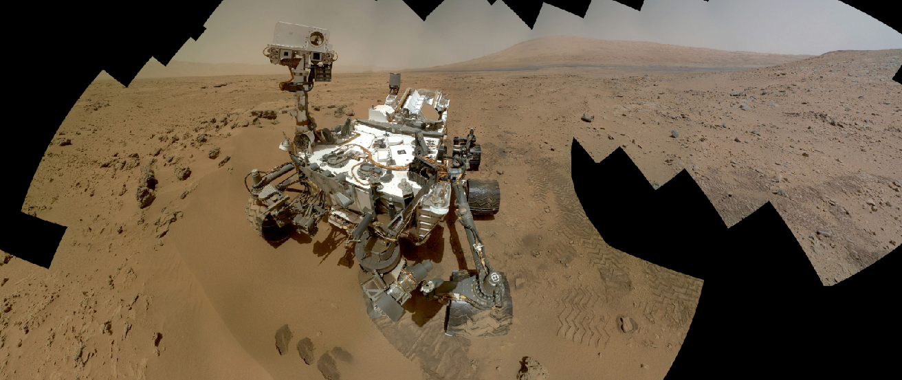 Image of the Curiosity Rover on the Martian surface. In this composite photograph we see the rover perched on the rusty-red Martian soil, with a series of hills and a dusty-colored sky in the background. The outline of this image is jagged due to the effects of combining the individual frames into a single montage.